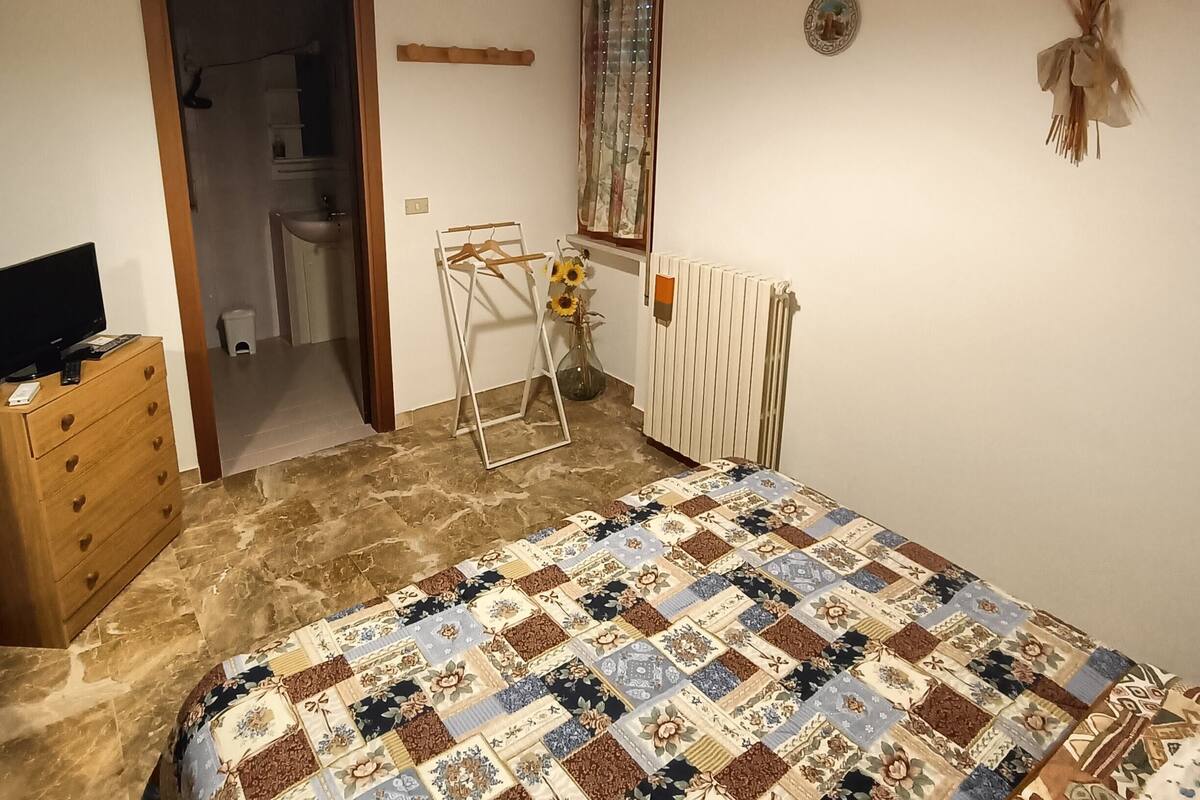 A bedroom with a quilted bedspread, a wooden dresser, a radiator, and a hanging plant.