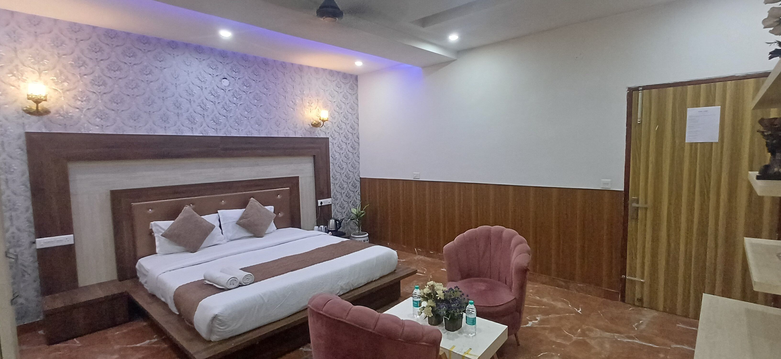 Habitación de hotel con cama, una silla, una mesita con un florero y botellas de agua, y una puerta de madera.