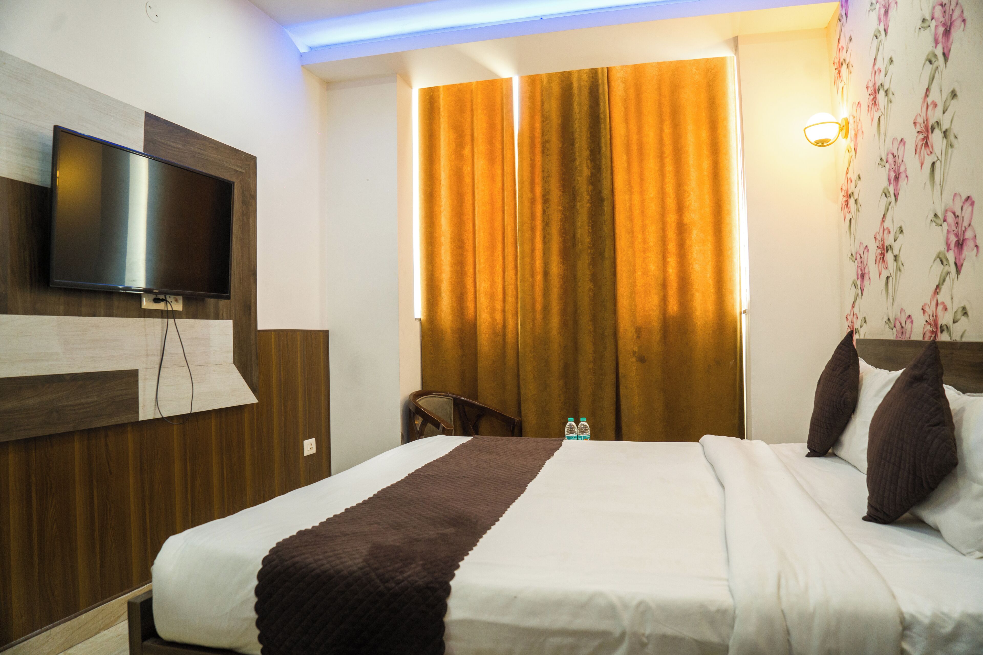 A hotel room with a bed, a flat-screen TV mounted on the wall, a wooden headboard, and a curtain.
