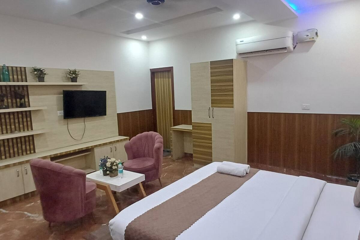 A hotel room with a bed, a TV, a small table with a vase, two chairs, a wardrobe, and an air conditioning unit.