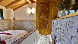 A rustic room with a stone wall, wooden ceiling, a bed with floral bedding, and a wall-mounted wooden artwork. - Stone House Only 150m From the sea (Umag)