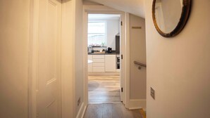 A hallway leading to a kitchen with white cabinets, a window, and a mirror on the wall. - Luxury Central London Apartment  Craven Street (London)