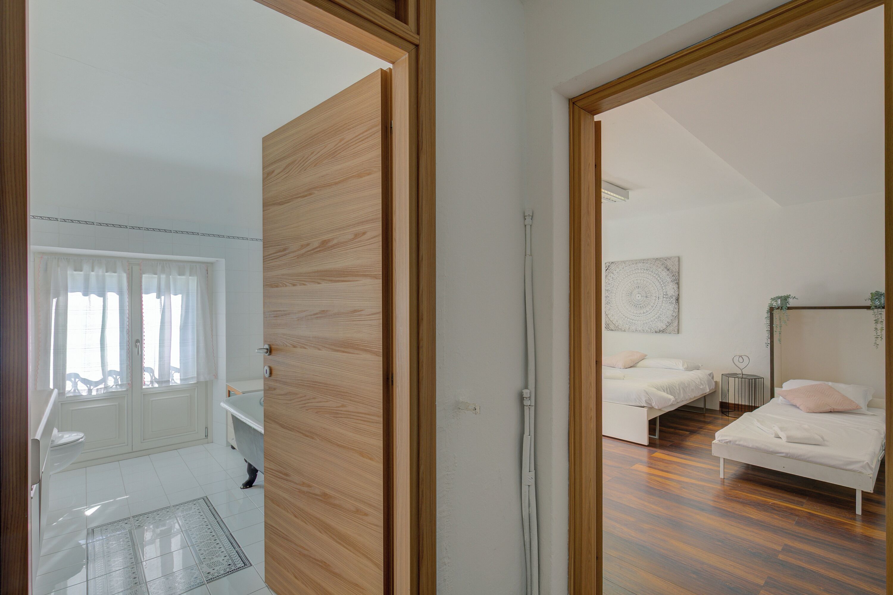 A room with a wooden door, a white sink, a window with curtains, a bed, and a wall-mounted artwork.
