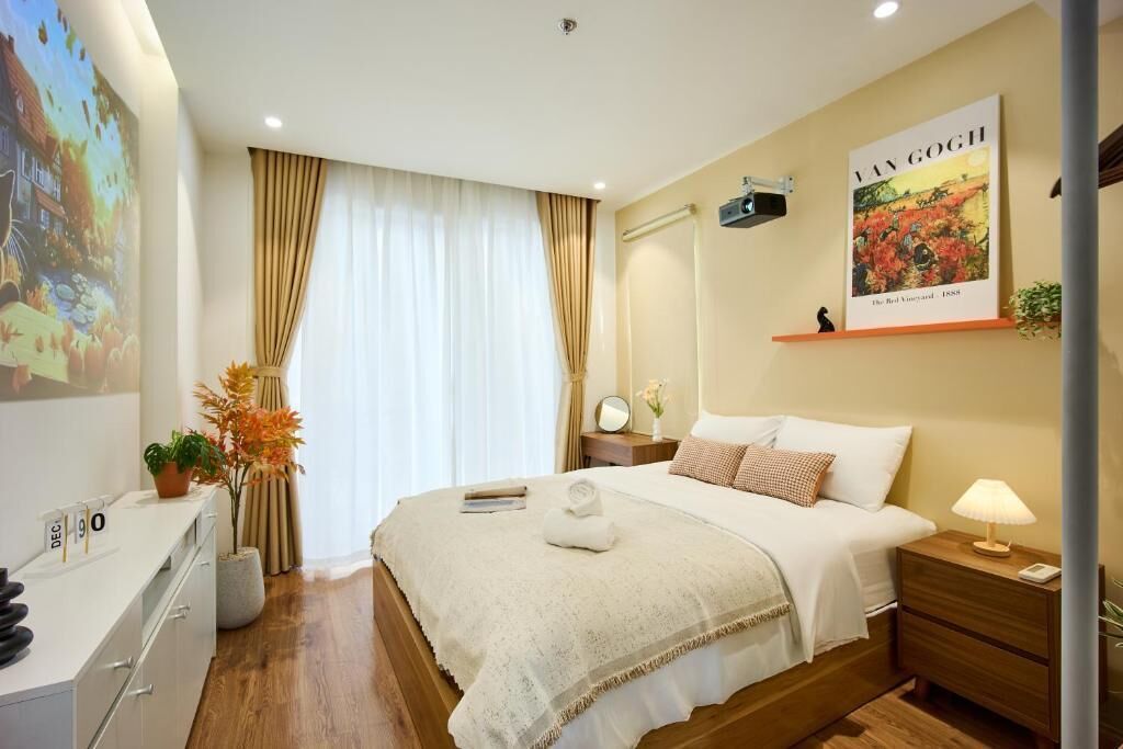 Une chambre d’hôtel comprenant un lit, une table de chevet, une commode, une fenêtre avec des rideaux, une œuvre d’art accrochée au mur et un bureau avec une plante.