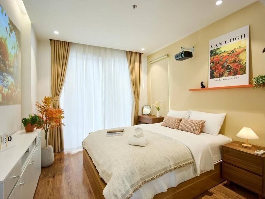 Une chambre d’hôtel comprenant un lit, une table de chevet, une commode, une fenêtre avec des rideaux, une œuvre d’art accrochée au mur et un bureau avec une plante.