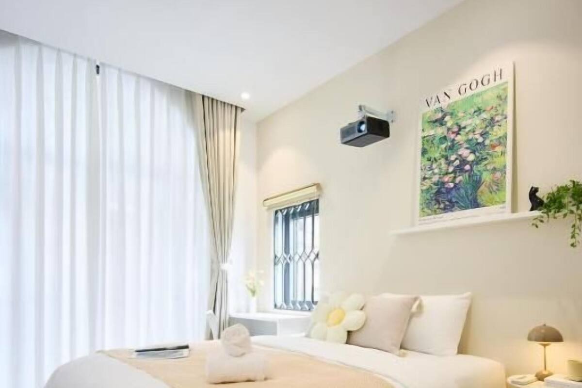 Una habitación de hotel con una cama grande, una ventana con cortinas, una obra de arte en la pared y una mesita de noche con lámpara.