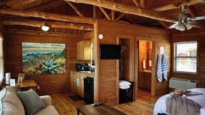 Interior de una cabaña con techo de madera, un cuadro de un paisaje montañoso, kitchenette, televisor, cama y ventana con vistas.