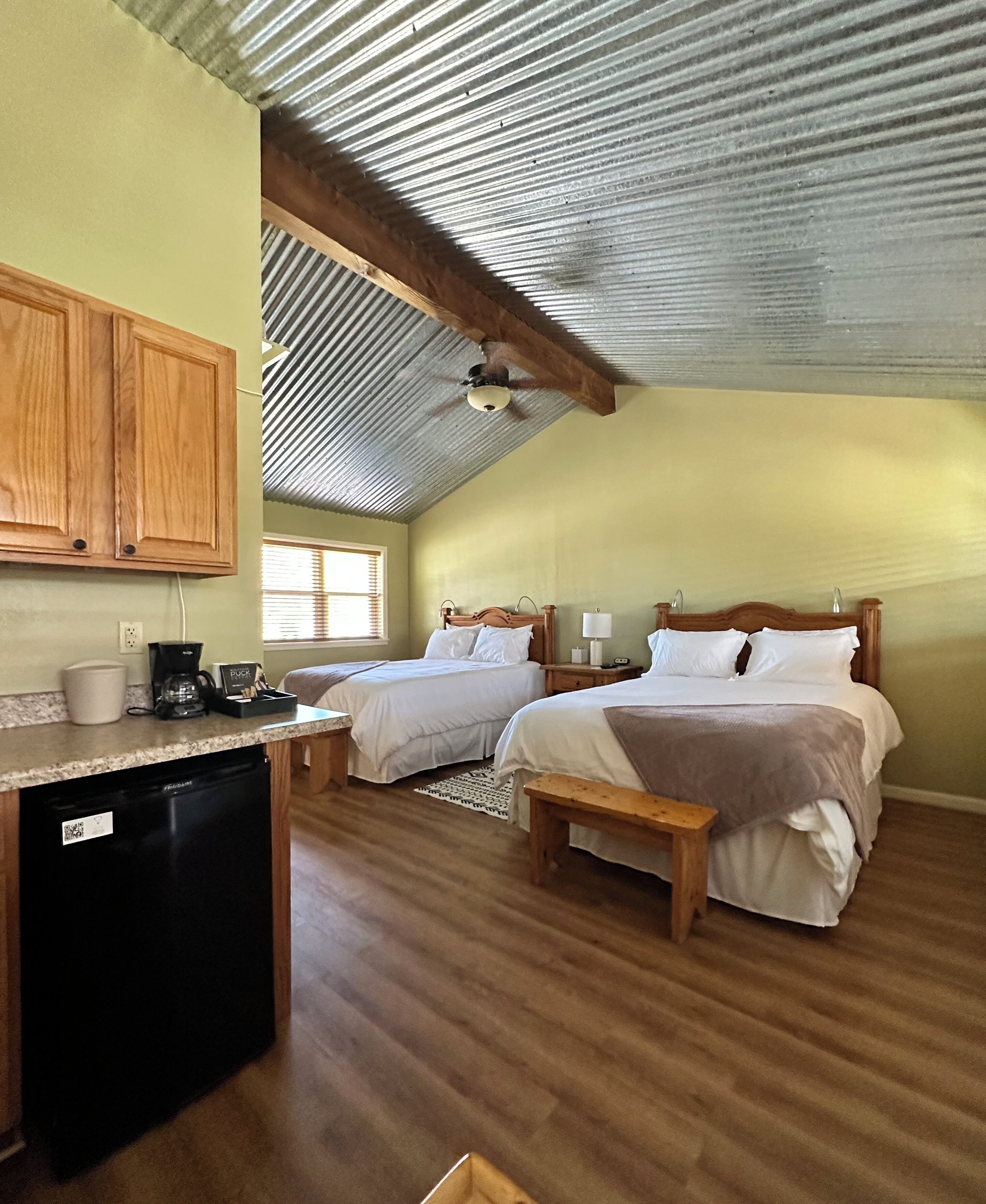 A room with two beds, a wooden bench, a small kitchen area with a dishwasher, and a ceiling fan.