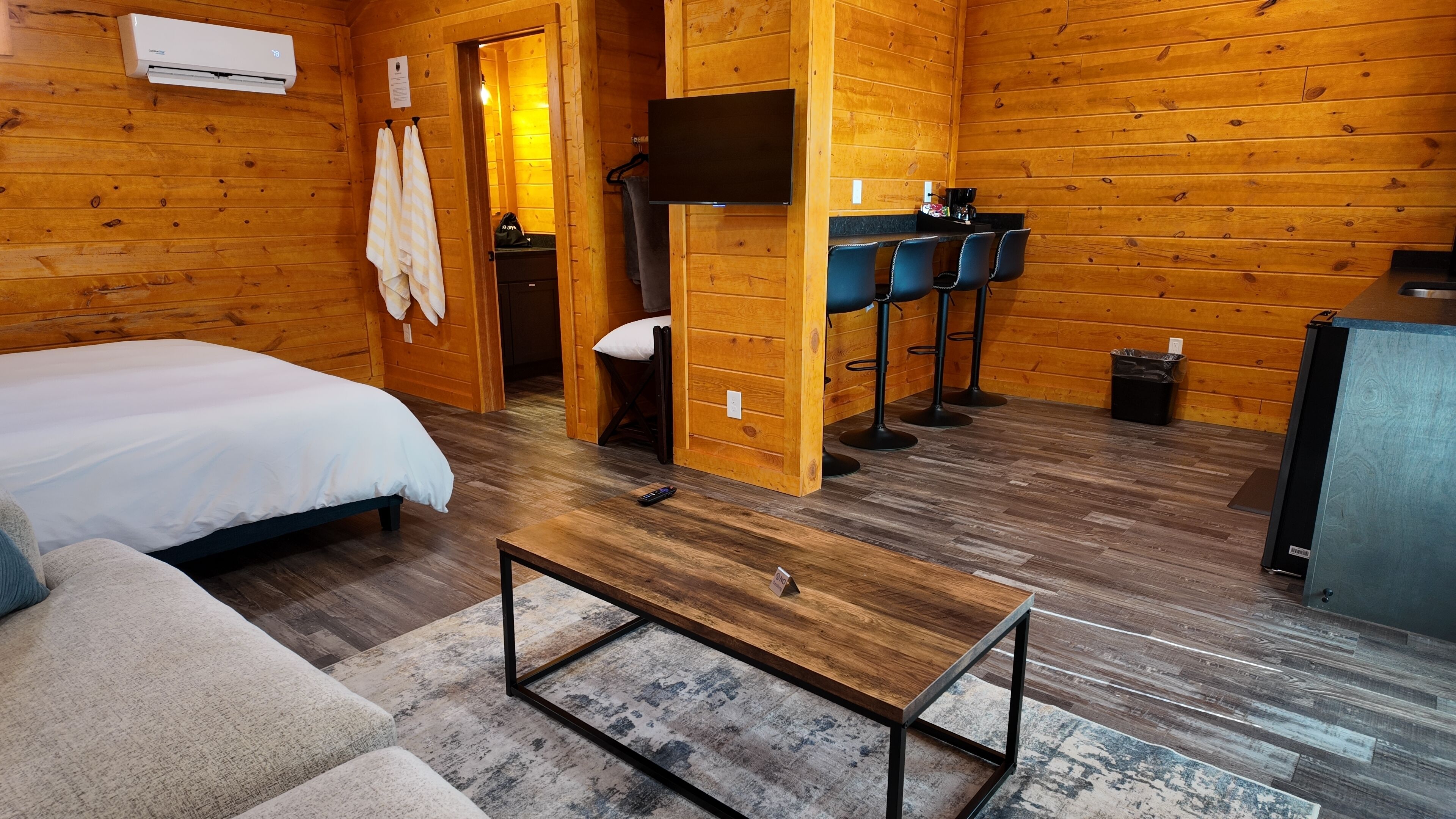 A wooden cabin room with a bed, a sofa, a coffee table, a TV, and a small kitchenette.