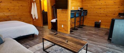A wooden cabin room with a bed, a sofa, a coffee table, a TV, and a small kitchenette.