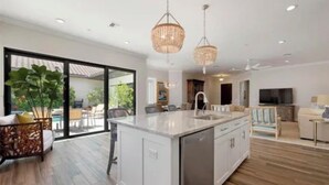 Une cuisine moderne dotée d’un grand îlot central, de luminaires suspendus et d’une porte-fenêtre coulissante donnant sur un espace extérieur.
