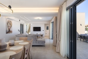 A modern living room with a sofa, TV, and dining area with placemats. - Villa Azure – Luxury Sea View Villa (Panormo)