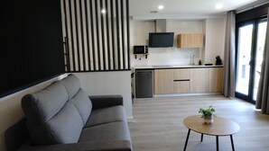 Ett modernt vardagsrum med en soffa, ett litet runt bord med en växt, en köksdel med en mikrovågsugn och en tv, samt en skjutdörr.