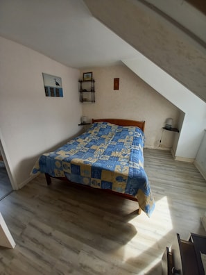 A bedroom with a sloped ceiling, a bed with a blue and yellow patterned bedspread, a small shelf with a picture, and a wall-mounted lamp. - Seaside fisherman's house Paimpol (paimpol)