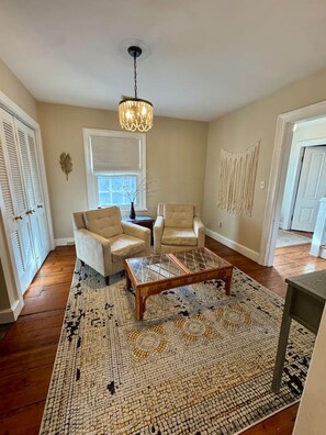 A room with a wooden floor, two beige armchairs, a coffee table, a hanging light fixture, a window with blinds, and a door. - Historic DT Lynchburg Apt, Walk To Shops & Dining (Lynchburg)