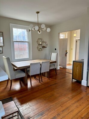 Un comedor con una mesa de madera, cuatro sillas, un adorno hexagonal en la pared, una lámpara en el techo y una puerta que da a otra habitación.