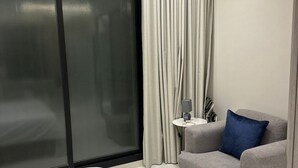Una habitación de hotel moderna con una cama, un sillón gris, una mesita con un altavoz y un gran ventanal con cortinas.