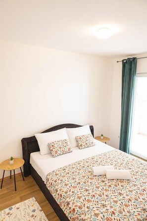A bedroom with a bed, two pillows, a bedside table with a plant, a small round table, a window with curtains, and a light fixture.