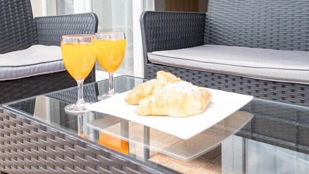 A glass-top table with two glasses of orange juice and a plate of croissants on a balcony with wicker furniture and a view of the sky. - Apartments Amare / Apartment 3 with Sea view (Neum)