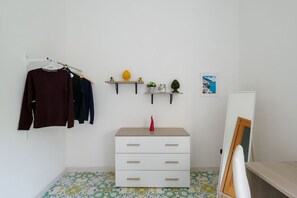 Ein Zimmer mit einem weißen Schminktisch, einem Kleiderständer mit verschiedenen Kleidungsstücken, zwei Wandregalen mit Dekoration und einem kleinen gerahmten Bild.