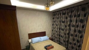 A small bedroom with a bed, a nightstand, a chair, a ceiling light fixture, and a window with curtains. - Stylish Downtown Cairo Apartment with Balcony View & Prime Location (Cairo)