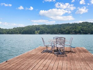 Een houten terras met een tafel en stoelen, uitkijkend op een rustig meer, omringd door een dicht bos.