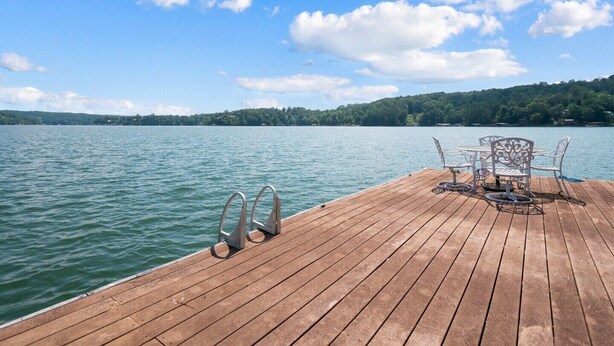 Um deck de madeira com guarda-corpos de metal, uma mesa e cadeiras de frente para um lago calmo com margem arborizada.