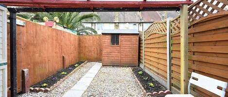 Een overdekt terras met een houten omheining, een klein houten gebouwtje en een bankje.