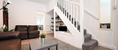 A living room with a brown leather sofa, a grey coffee table, a staircase with white railings, and a TV on the landing.