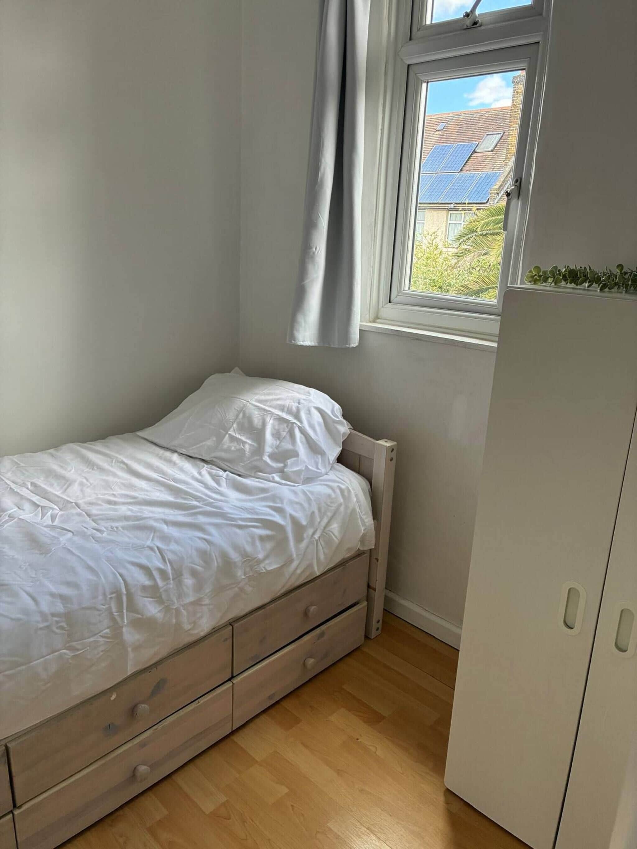 Een kleine kamer met een enkel bed, raam, houten vloer en een witte kast.