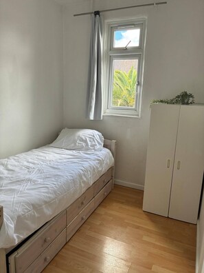 Une petite chambre avec un lit simple, un plancher en bois, une fenêtre donnant sur un espace verdoyant et une armoire blanche.