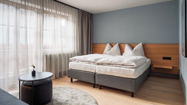 A modern bedroom with a bed, a round side table, and a large window with sheer curtains. - Family room Markus in Wagrain up to 3 pers - Julius Appartements (Hofmark)