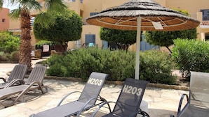 Un espace aménagé au bord de la piscine, avec des chaises longues, un palmier et un bâtiment doté de balcons en arrière-plan.