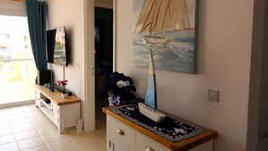 Une pièce avec un téléviseur posé sur un meuble, un tableau représentant un voilier, un buffet et une porte donnant sur une autre pièce.