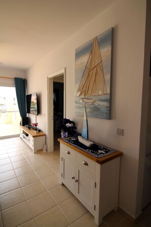 A room with a TV on a stand, a painting of a sailboat, a cabinet, and a door leading to another room. - STAY Limnaria Gardens Apartment (Paphos)