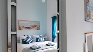 Une chambre avec un grand miroir, un lit avec une literie bleue et blanche, un tableau accroché au mur et une table de chevet en bois.