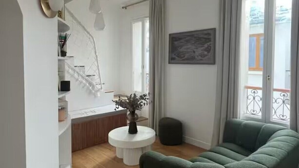 Una sala de estar con un sofá verde, una mesa redonda blanca con un florero, un cuadro en la pared y un balcón con barandilla.