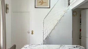 Una habitación con una mesa de mármol, una puerta blanca, un cuadro enmarcado y una escalera blanca.