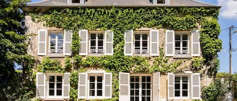A two-story building with white shutters, a stone facade, and a garden at the front.