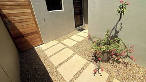 Un pati amb una porta de fusta, una planta en test amb flors roses i un camí empedrat.