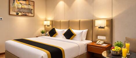 Habitación de hotel con una cama grande, mesitas de noche, una pequeña mesa con fruta y bebidas, y un cuadro en la pared.