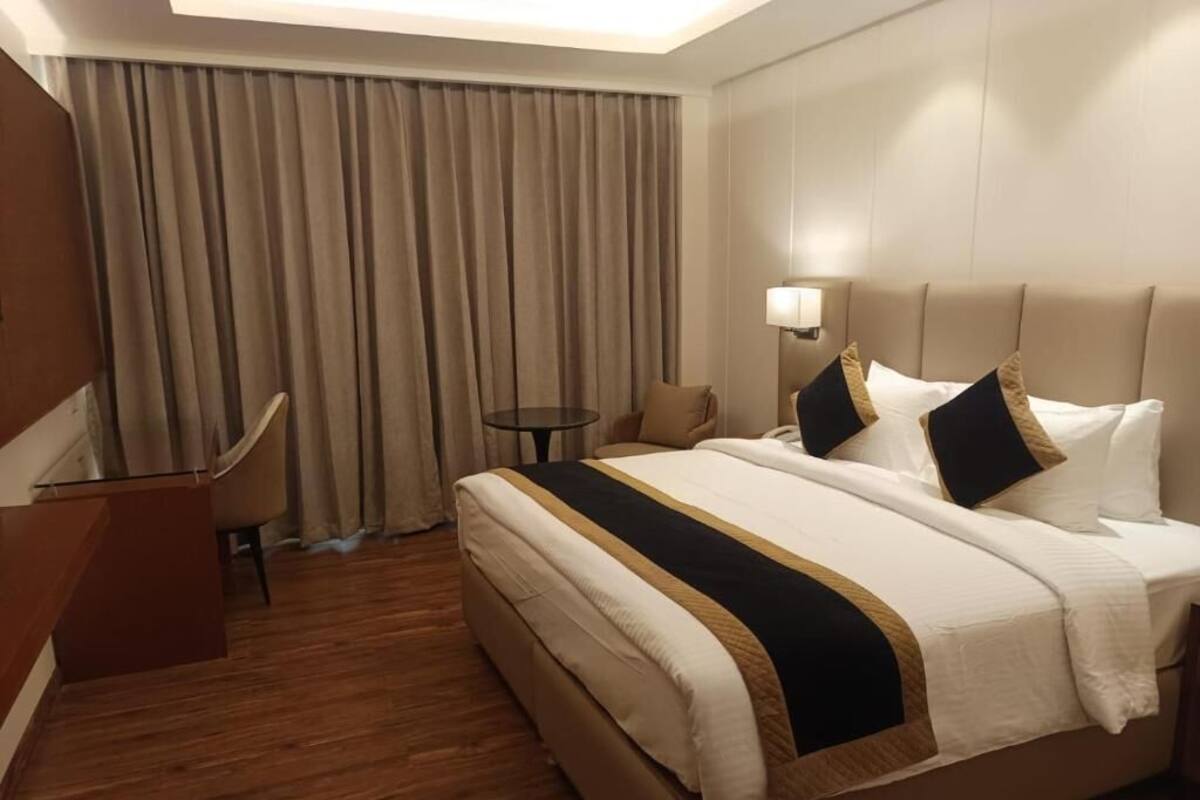 Habitación de hotel con cama, escritorio, silla, lámpara y vistas al exterior a través de cortinas.