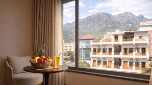 A room with a view of mountains, a table with fruit and a drink, a chair, and a window with curtains. - One Earth Heritage Katra (Katra)