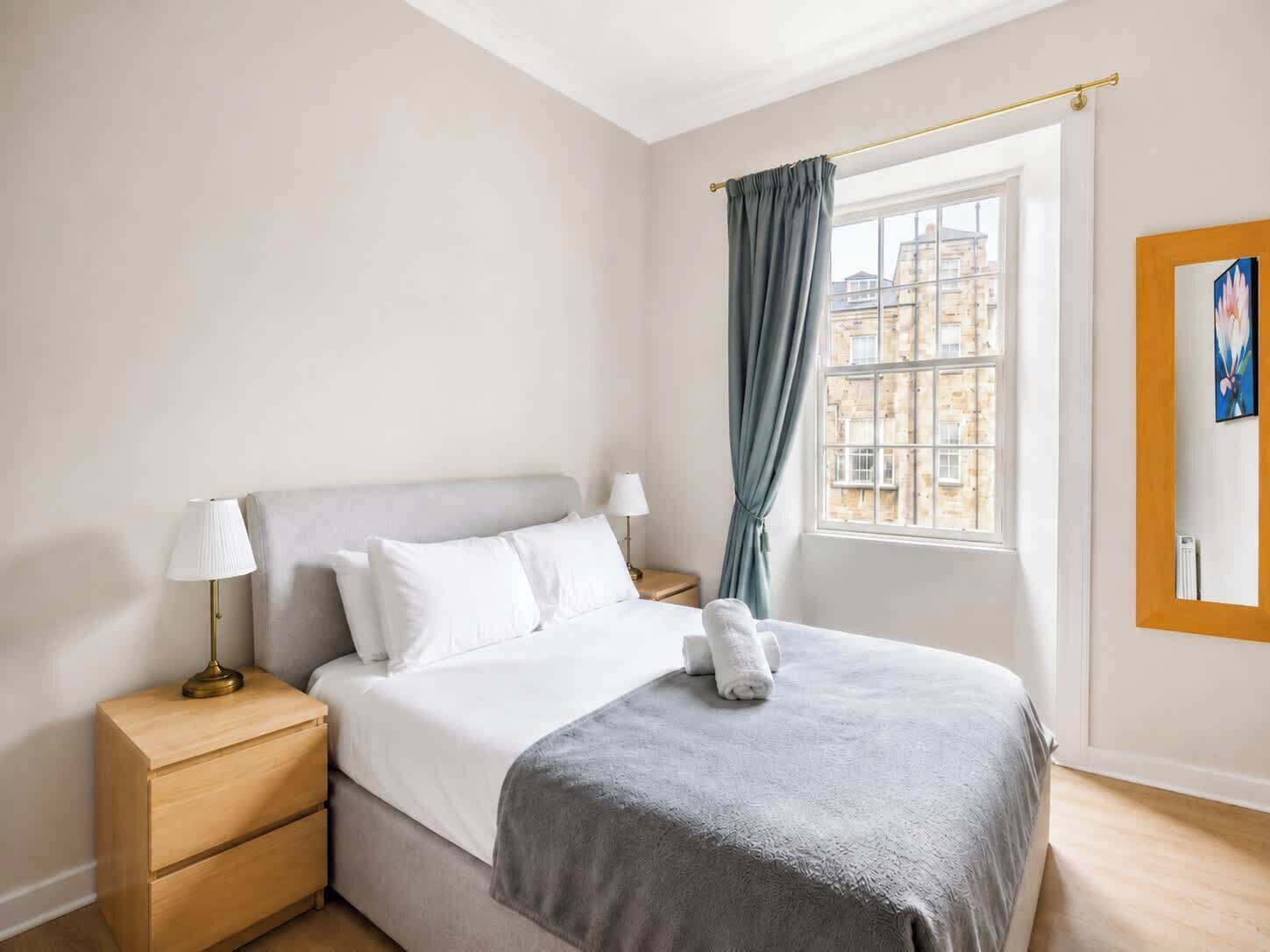 A hotel room with a bed, bedside tables, a window with curtains, and a painting on the wall.