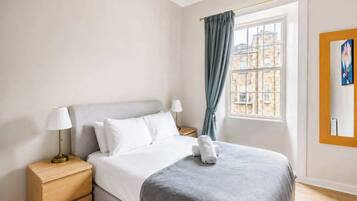 A hotel room with a bed, bedside tables, a window with curtains, and a painting on the wall.