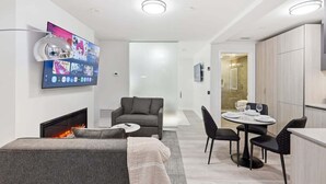 Ein modernes Wohnzimmer mit Kamin, einem Wand-Fernseher, einem grauen Sofa und einem Essbereich mit Tisch und Stühlen.