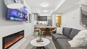 Un salon moderne avec une cheminée, un téléviseur à écran plat fixé au mur, un coin repas avec une cuisine en arrière-plan et un canapé gris agrémenté de coussins.
