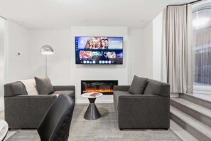 Ett modernt vardagsrum med två grå soffor, en platt-TV som är monterad på väggen, en öppen spis och ett stort fönster med gardiner.