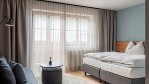A modern hotel room with a bed, sofa, round table, and large windows with sheer curtains. - Room Leni with balcony - Julius Appartements Wagrain, 22 m² up to 3 persons (Hofmark)