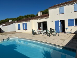 Ein Poolbereich mit Tisch und Stühlen, eine überdachte Terrasse und ein Gebäude mit blauen Fensterläden.